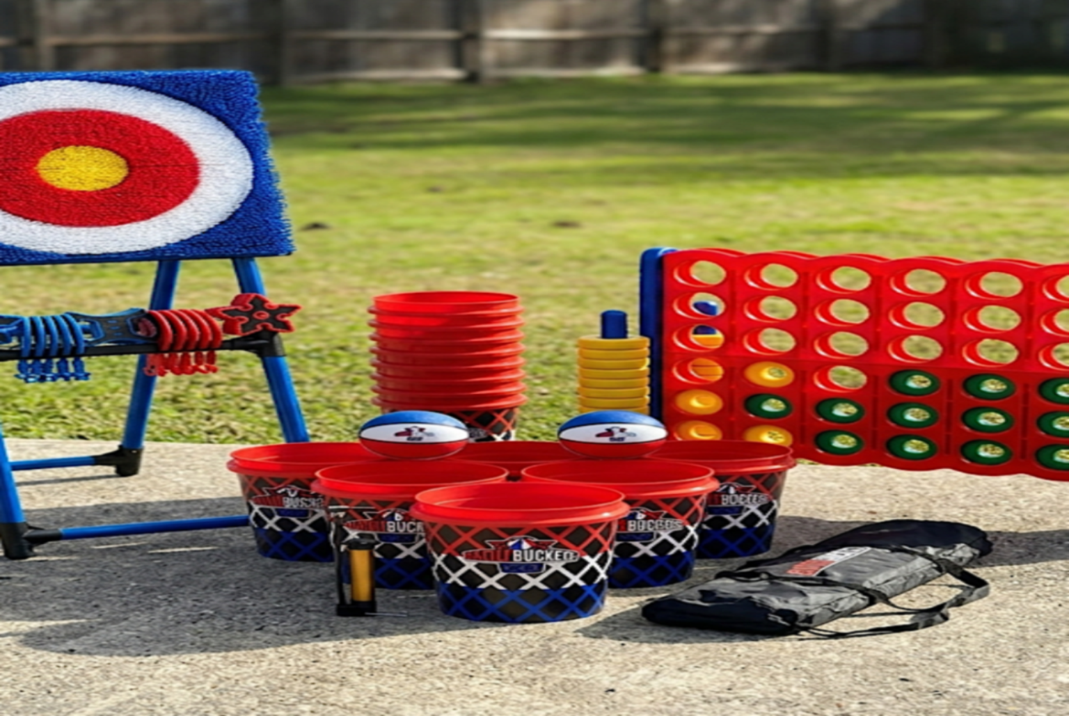 Outdoor games including a target, buckets, and a Connect Four game set on a concrete surface with grass in the background.