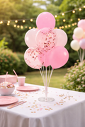 Decorative pink balloons with confetti on a table outdoors with string lights and greenery.